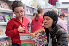 An adult and child in red read a "World of Reading" book in a library. Shelves with colorful books in the blurred background. Cozy mood.